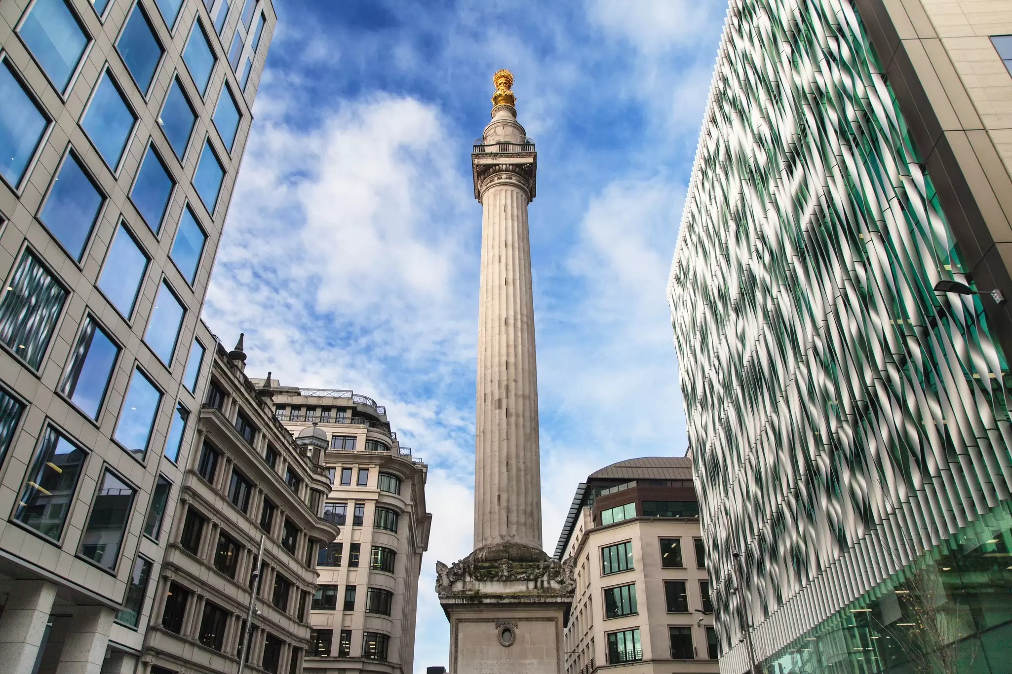 A tall stone column - Monument to the Great Fire of London - with a gold flame on the top stands among modern buildings