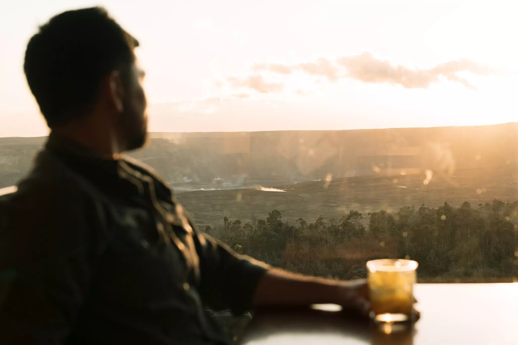 A man looks out a window at sunset while he sits at a table with a cocktail