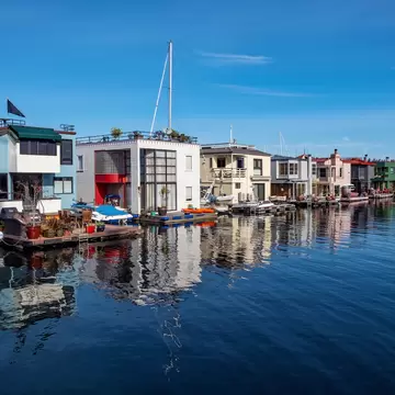 Row of blue, white, cream, pink and green houseboats moored on calm deep-blue water under a clear sky, a sailboat mast behind