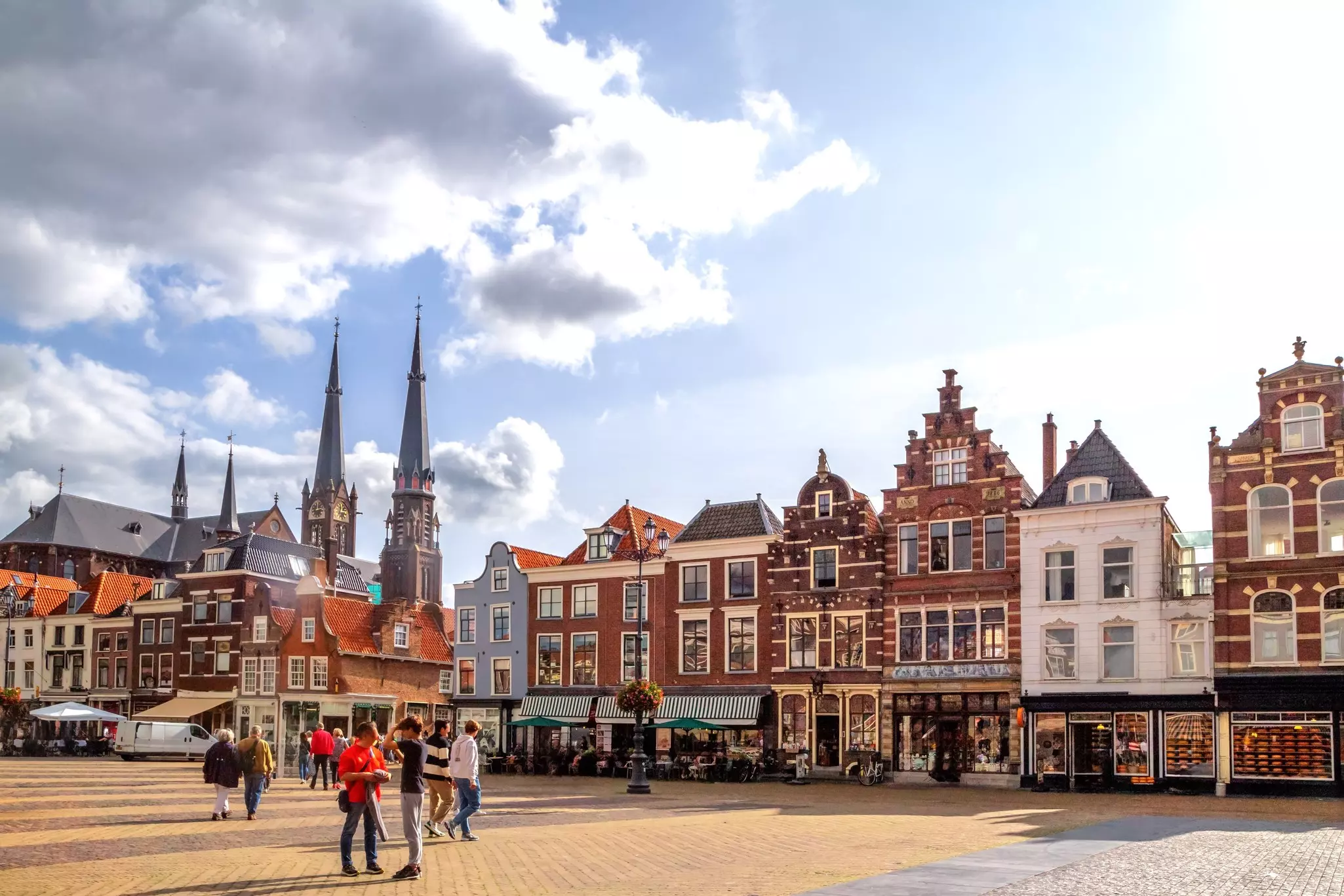 People wander through a medieval market square lined with shops and cafes. A tall church with two steeples stands beyond.