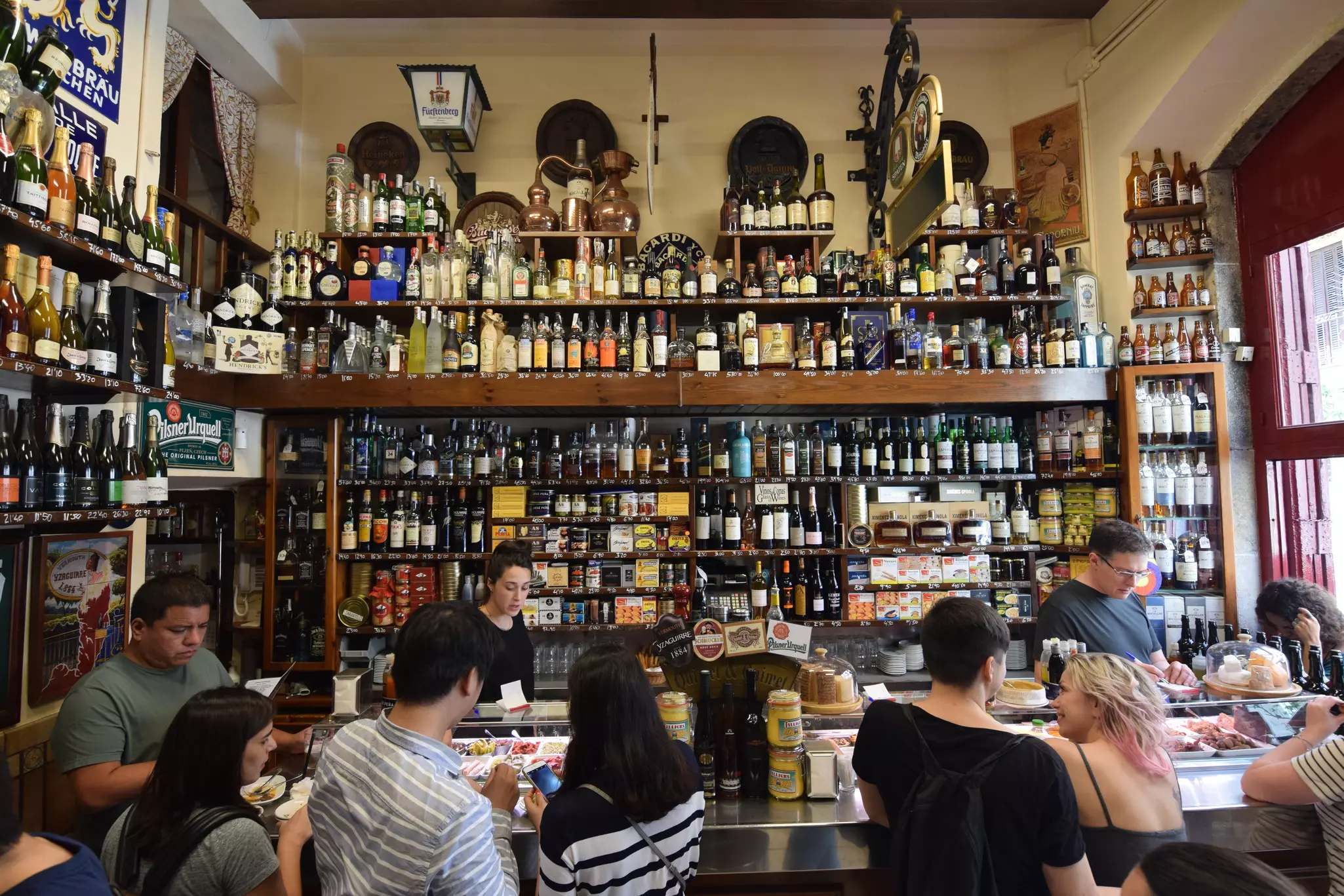 People eating and drinking in a tapas bar with a large selection of alcoholic drinks on the wall