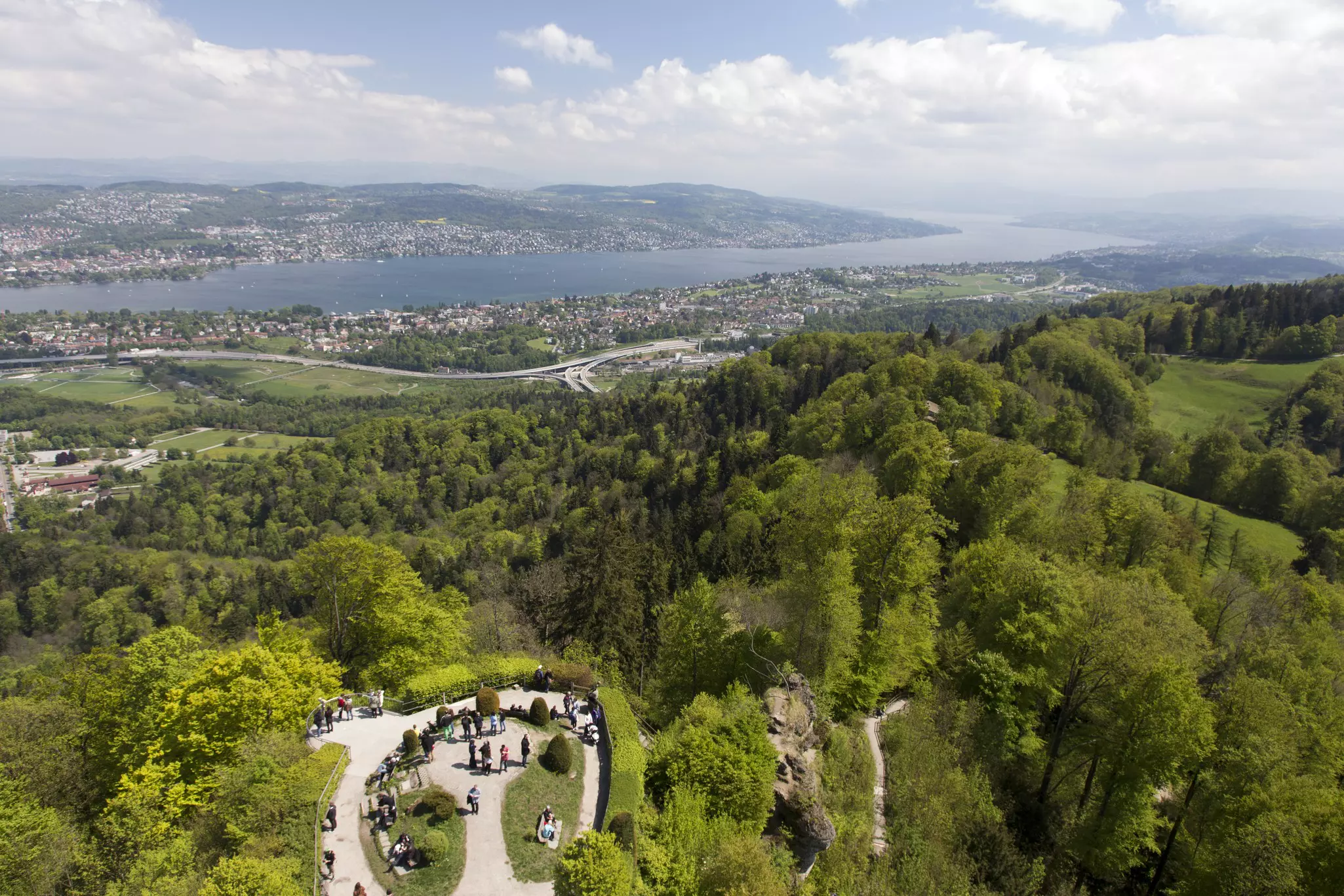 An aerial view of a hilltop covered with trees. People are seen at a viewpoint on the hilltop, which overlooks a city and lake below.