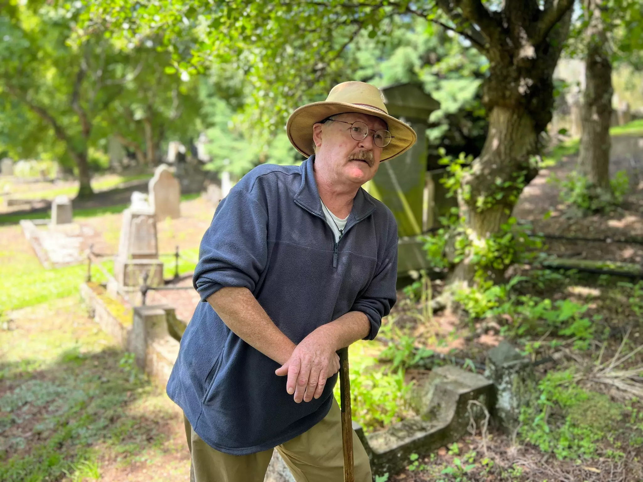 A man leans on a walking stick in a graveyard as he tells stories.