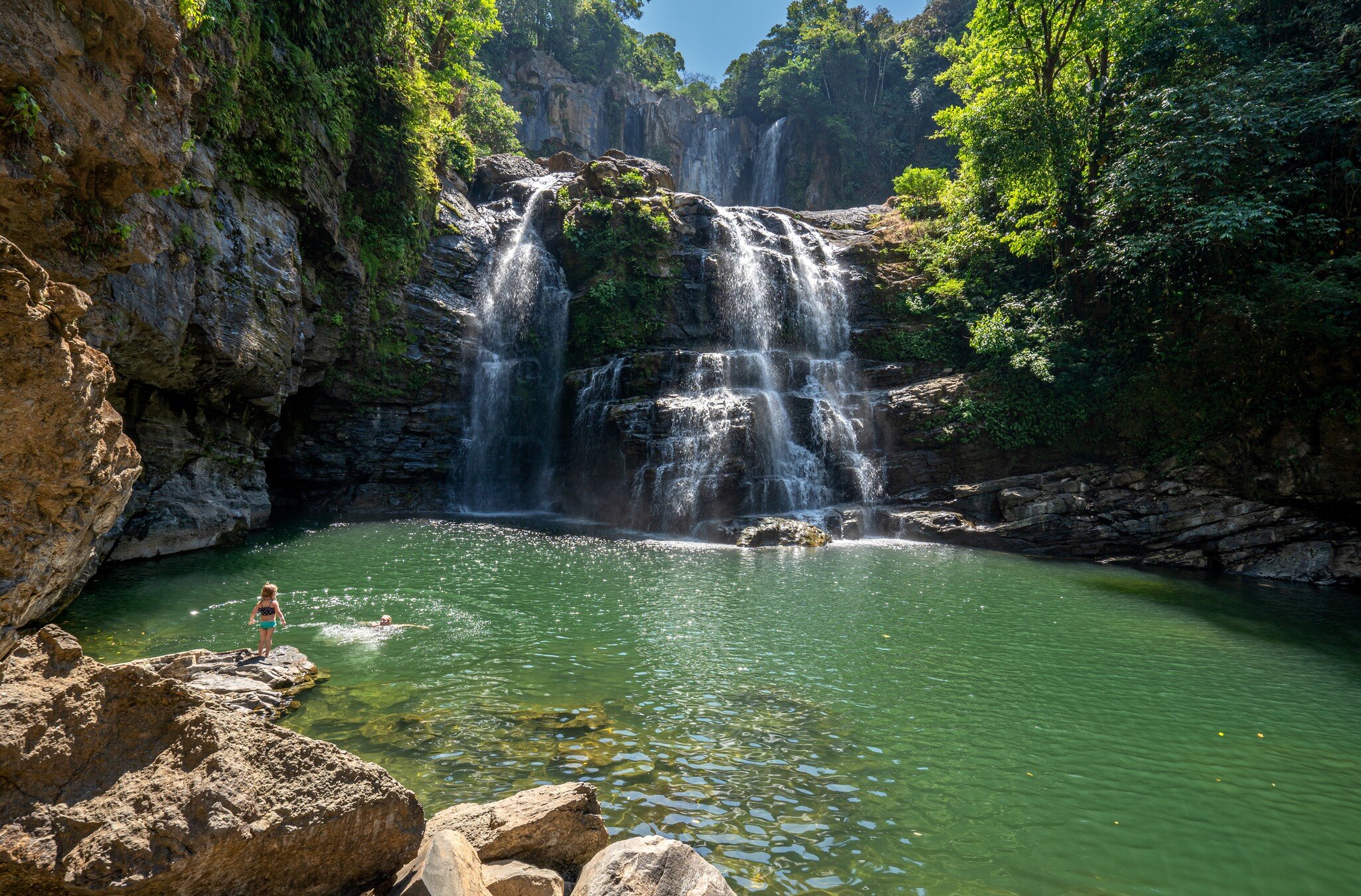 Two-tier waterfall spilling over dark rock ledges into a green pool ringed by boulders and forest, with a few swimmers.
