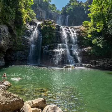 Two-tier waterfall spilling over dark rock ledges into a green pool ringed by boulders and forest, with a few swimmers.