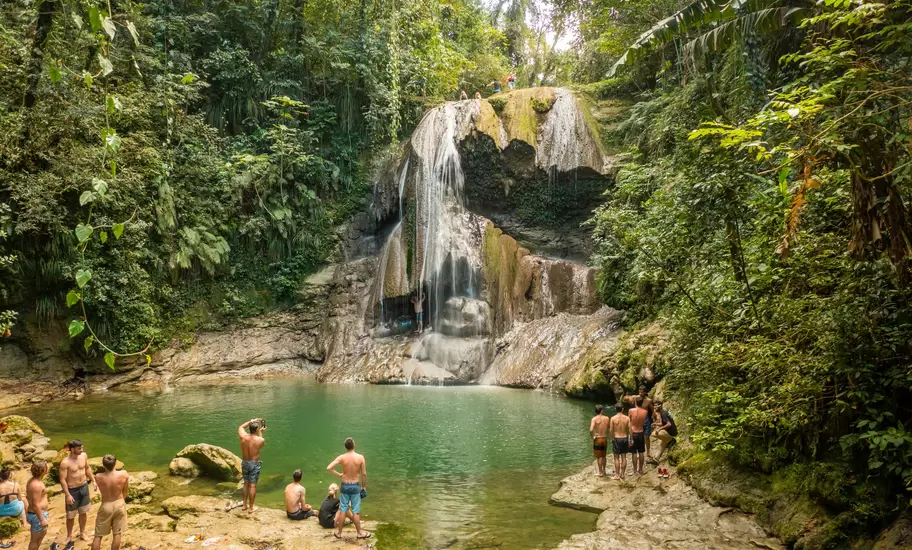 Waterfall spilling into a green pool in Puerto Rico's rainforest, with swimmers gathered on the rocky banks