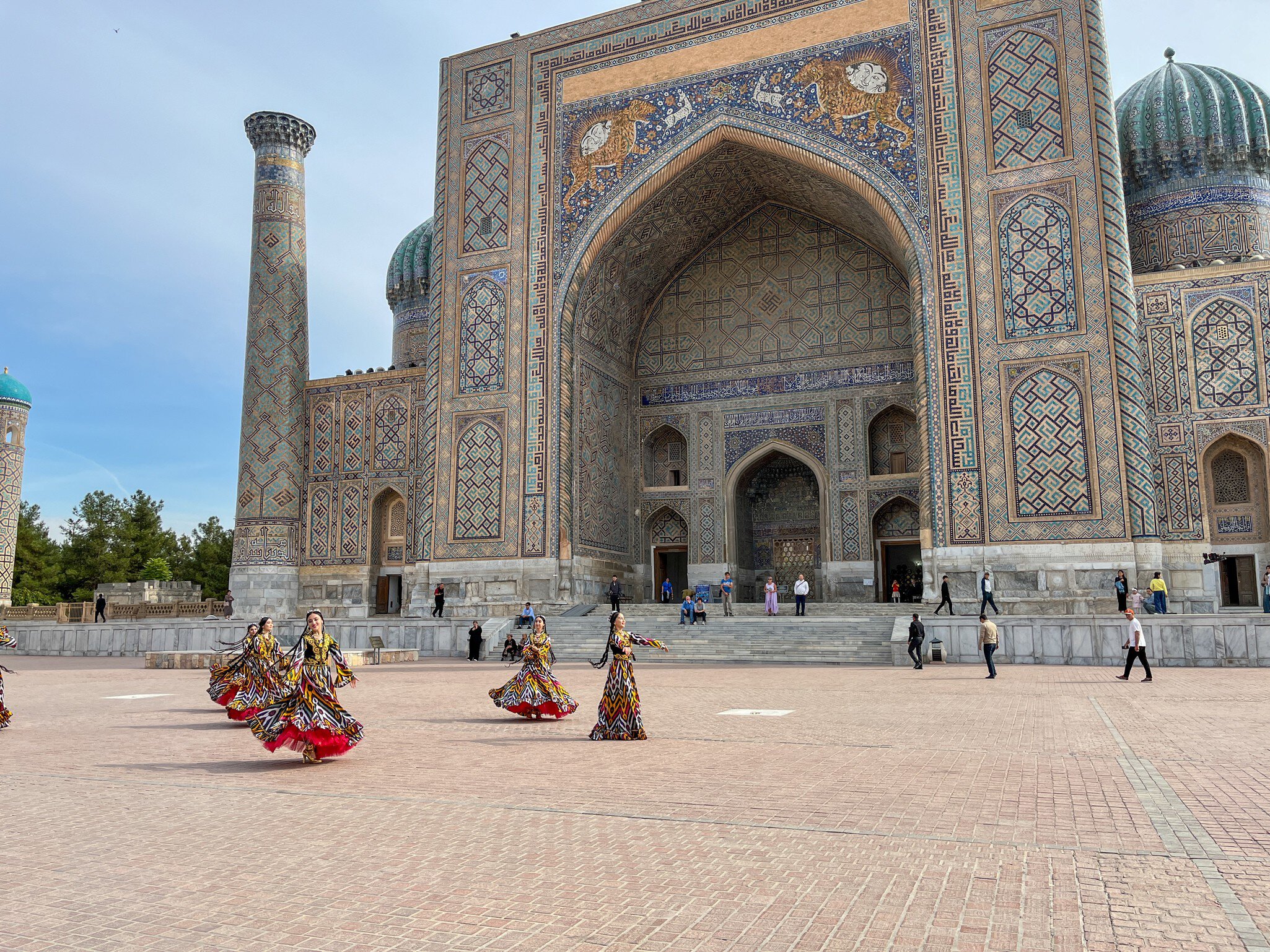 Sher-Dor Madrasah, Samarkand: tiled portal with tiger-and-sun mosaic, minaret and ribbed domes; women in ikat dresses dance