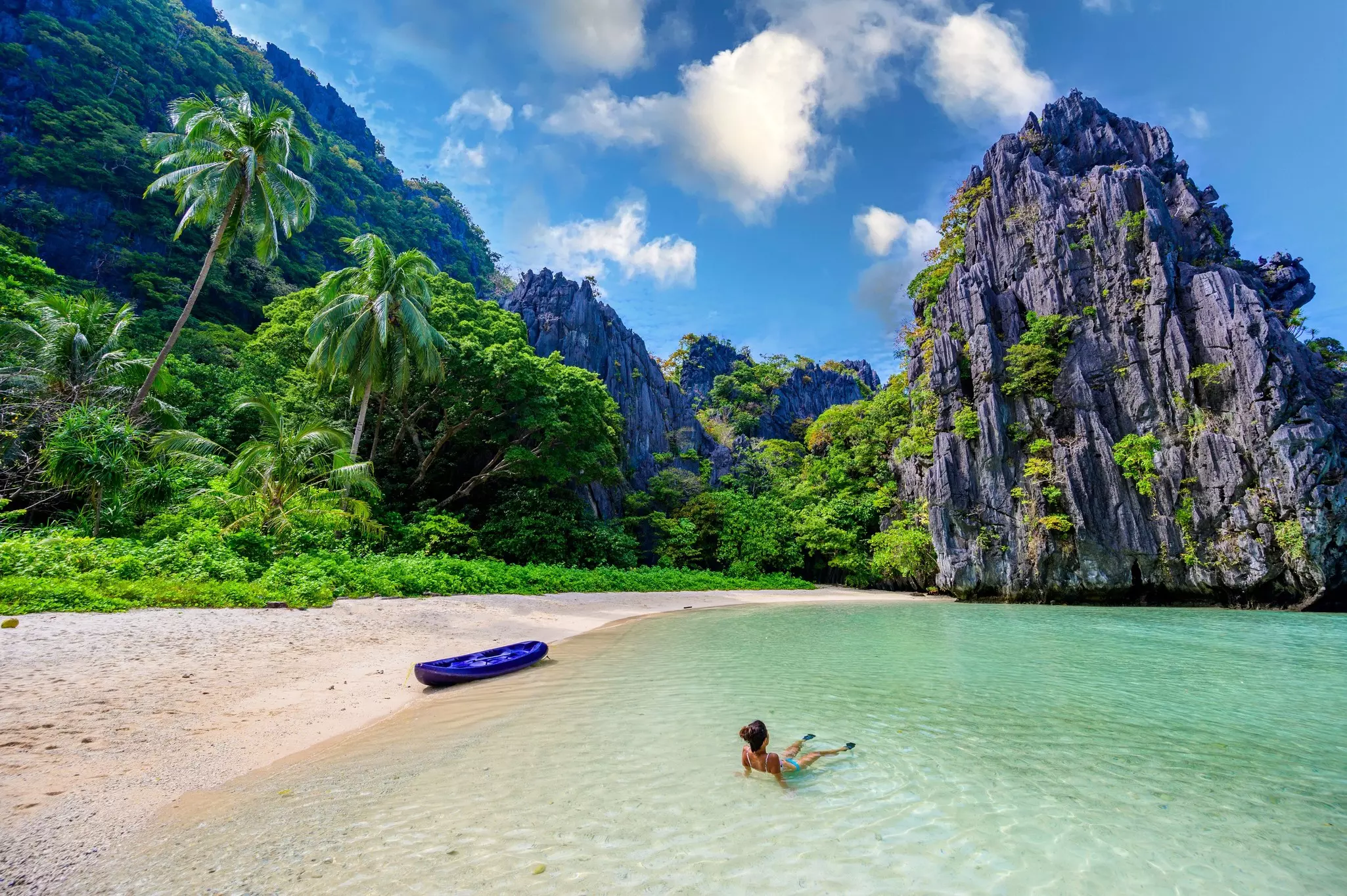A woman floats in the shallow waters at a tropical beach. She is alone; karst towers and palm trees are available on the shore
