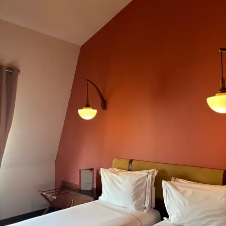 A two-piece bed with a white covering and pillows against a red wall with a lamp sconce.