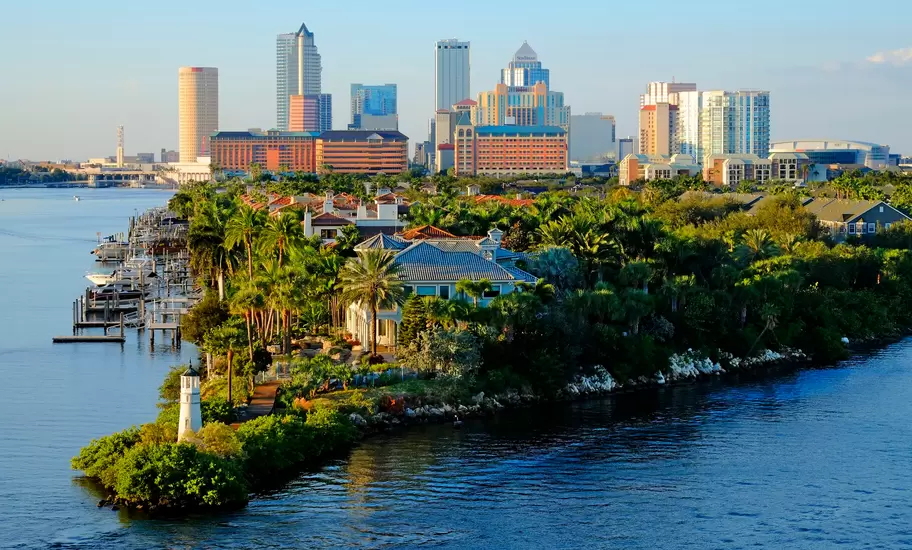 Waterfront view of a downtown skyline behind a lush green point dotted with luxury homes, docked boats and a small lighthouse