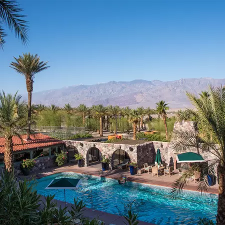 A rectangular pool enclosed by lounge chairs and umbrellas and a low stone building. Palm trees rise in the background.