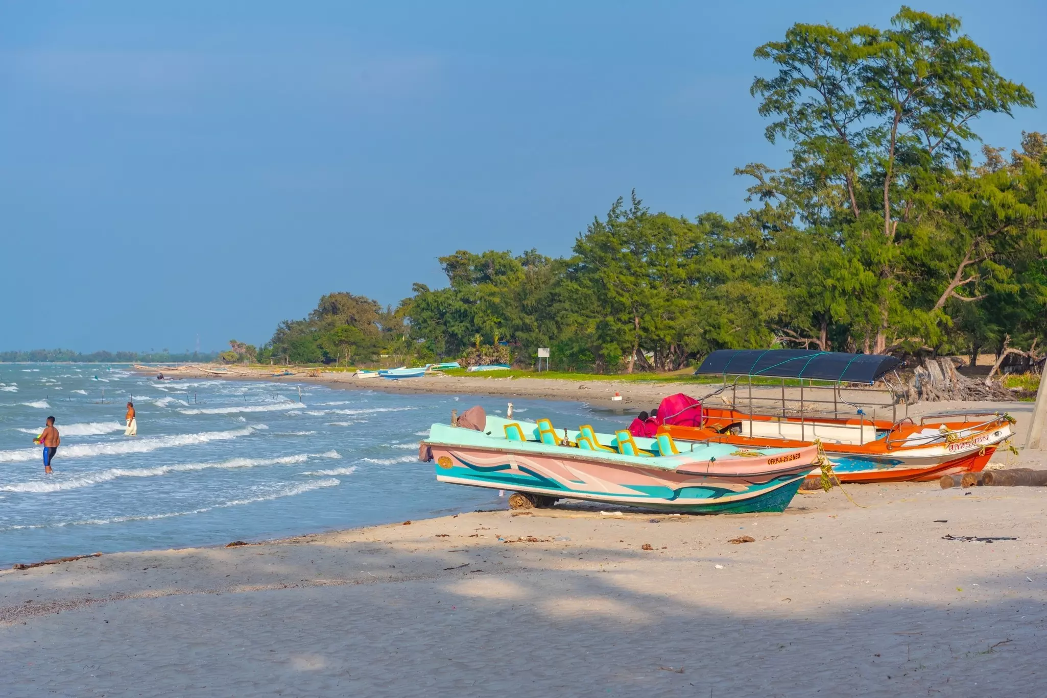 Colorful fishing boats are parked on the sand at a tropical beach. People are seen wading into the water, and lush vegetation covers the coast just in from the beach.