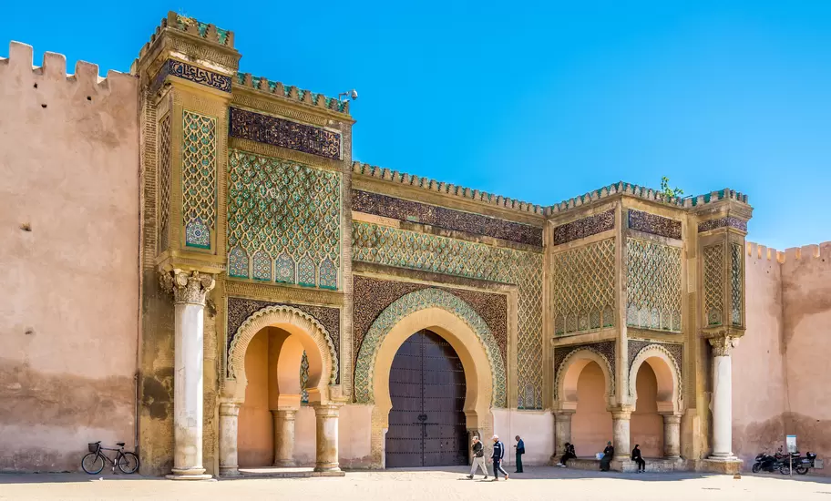 An ornate tiled Moroccan city gate with a large horseshoe arch and marble columns under a clear blue sky