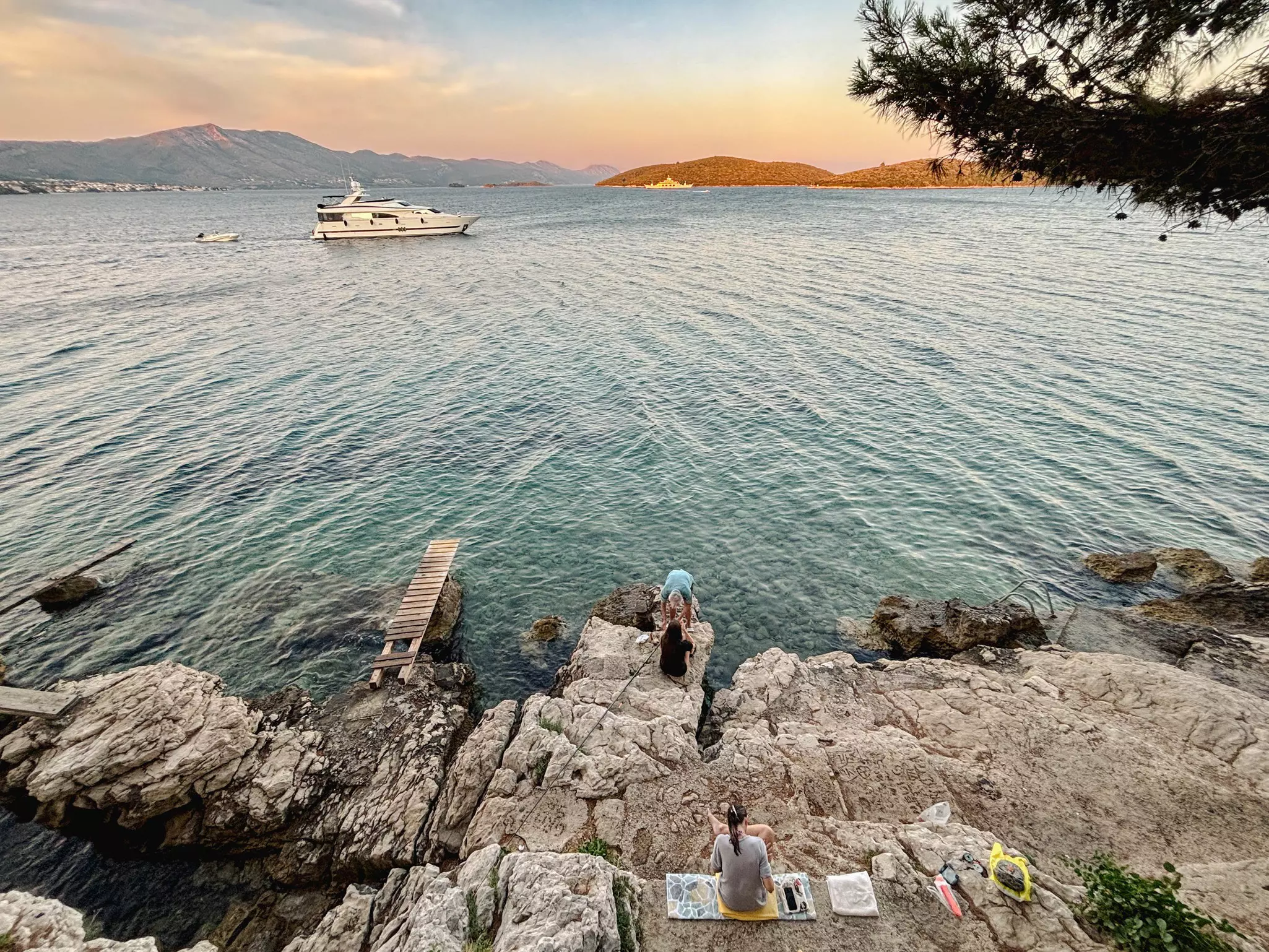 People sit on rocks by the sea; a boat is offshore.