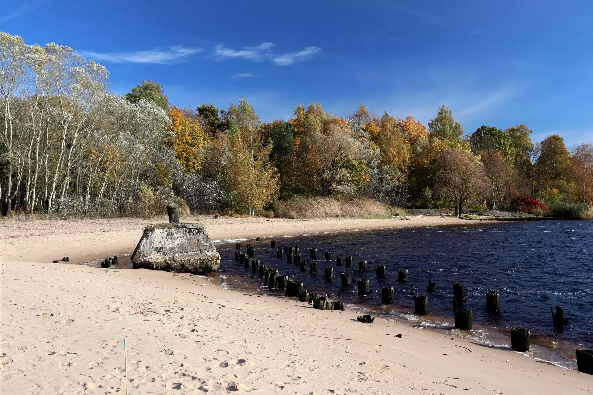 A rock and other barriers lie at the water line of a beach, under a blue sky
