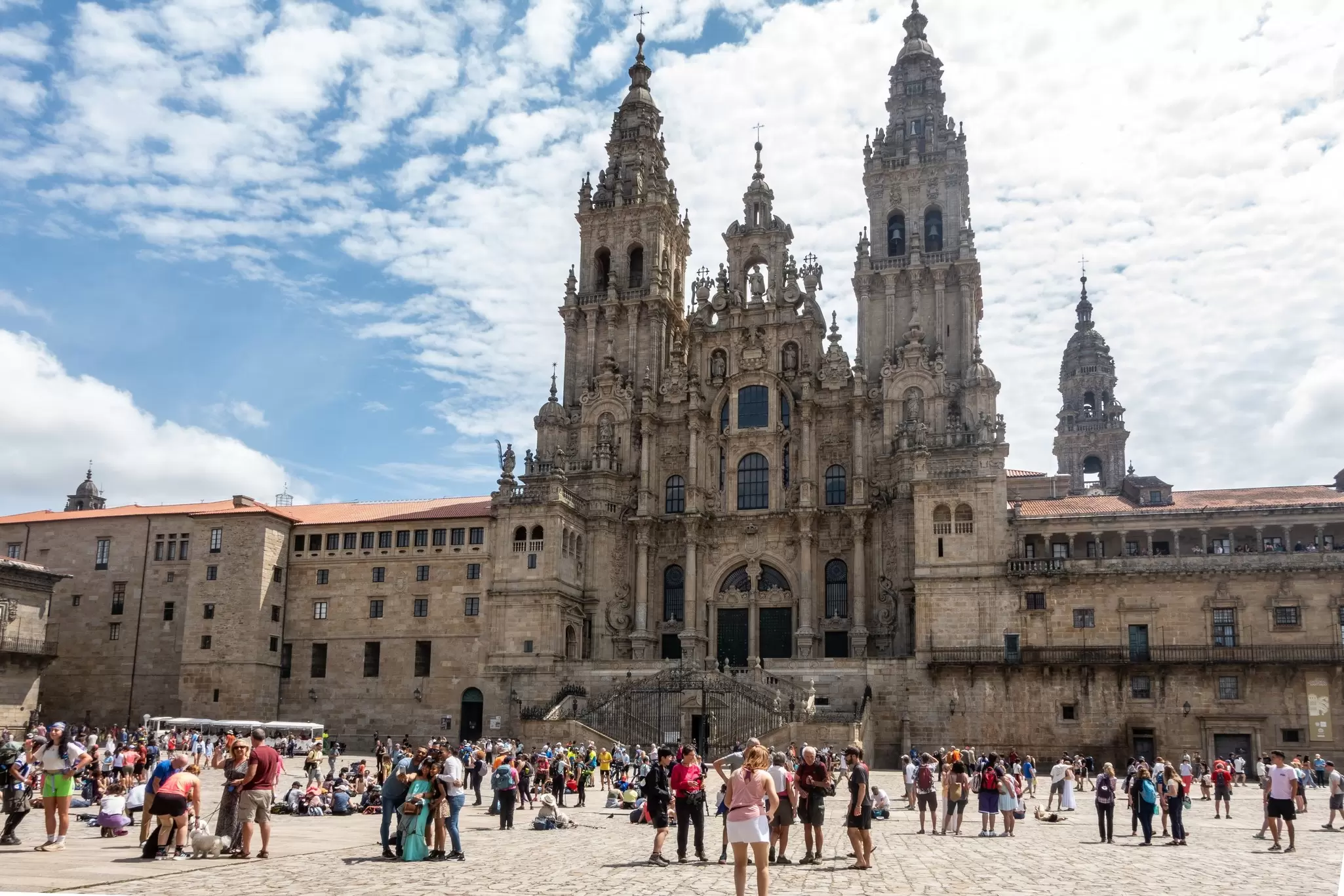 Pilgrims and tourists exploring the bustling square in front of the magnificent Catedral de Santiago de Compostela under a partly cloudy sky in Spain.