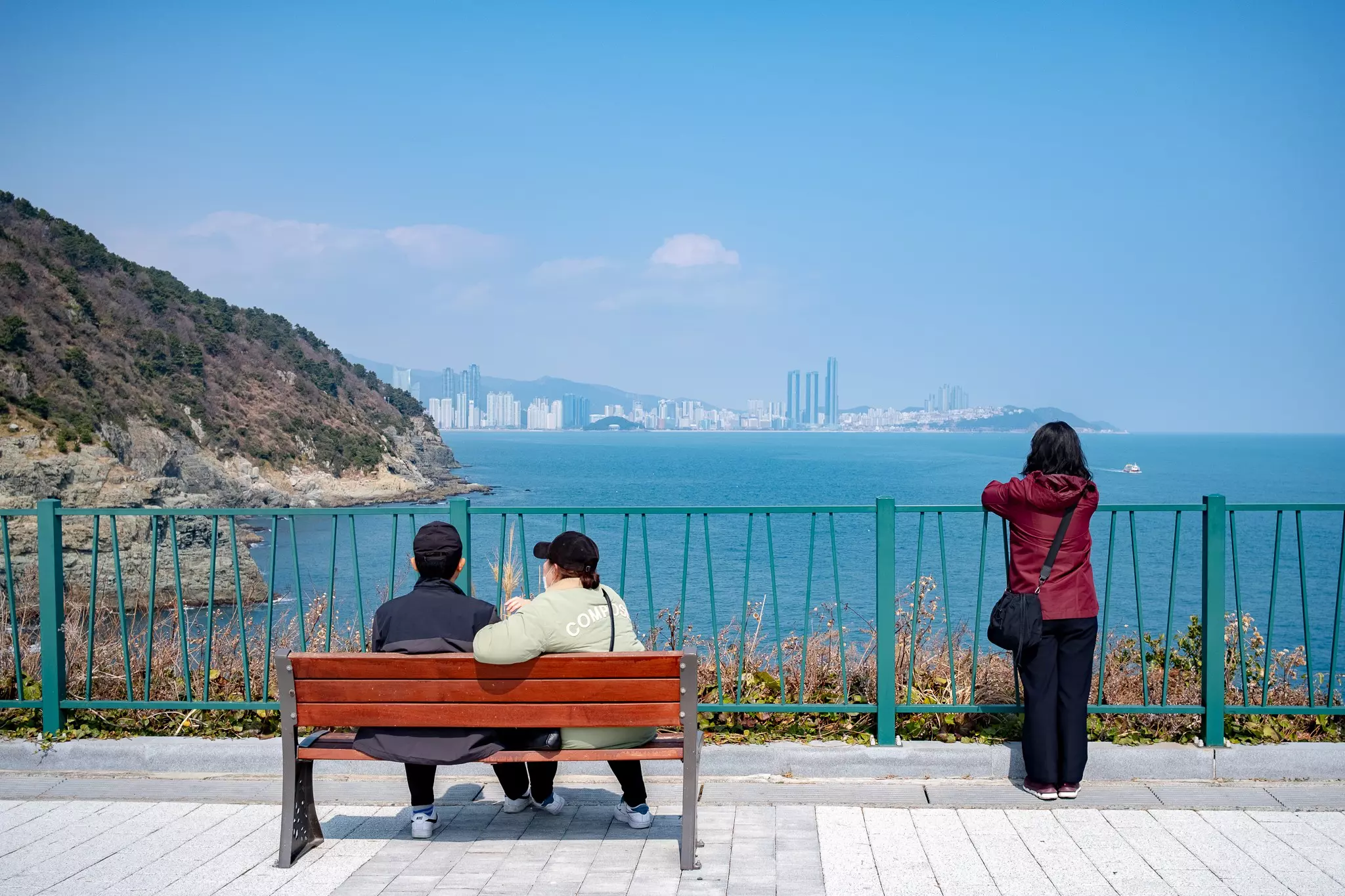 Two people sit on a bench and one stands at a railing looking out over the coastal scenery and towards a high-rise city on the Igidae Coastal Trail in Busan