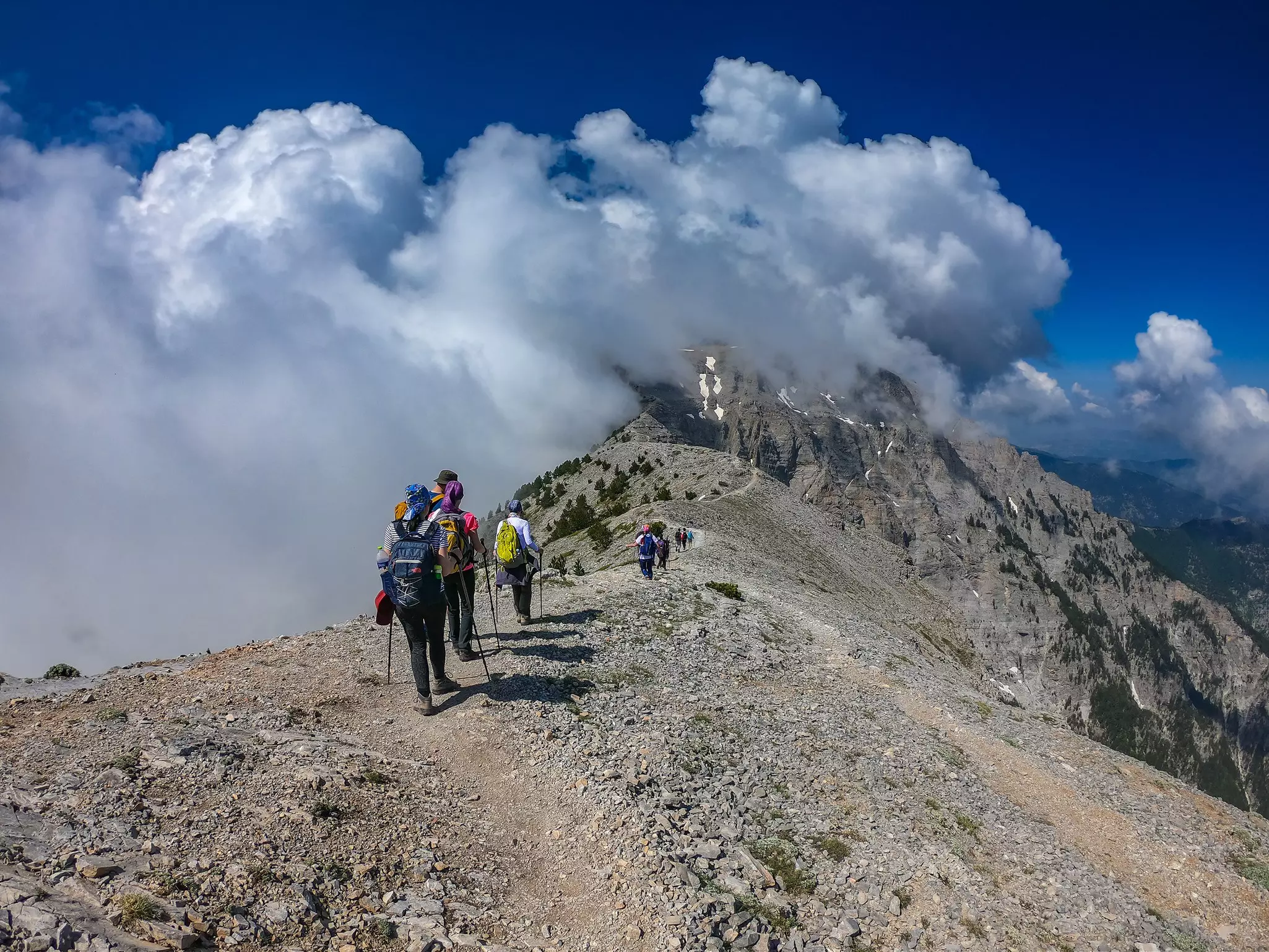 Hikers with backpacks and walking sticks follow a stony path up a vast mountain towards a snow-capped peak.