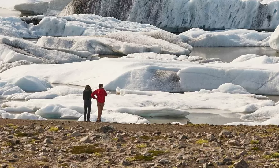 Two people on the shore below icebergs and the crevassed Fjallsjökull glacier, Vatnajökull National Park, Iceland.