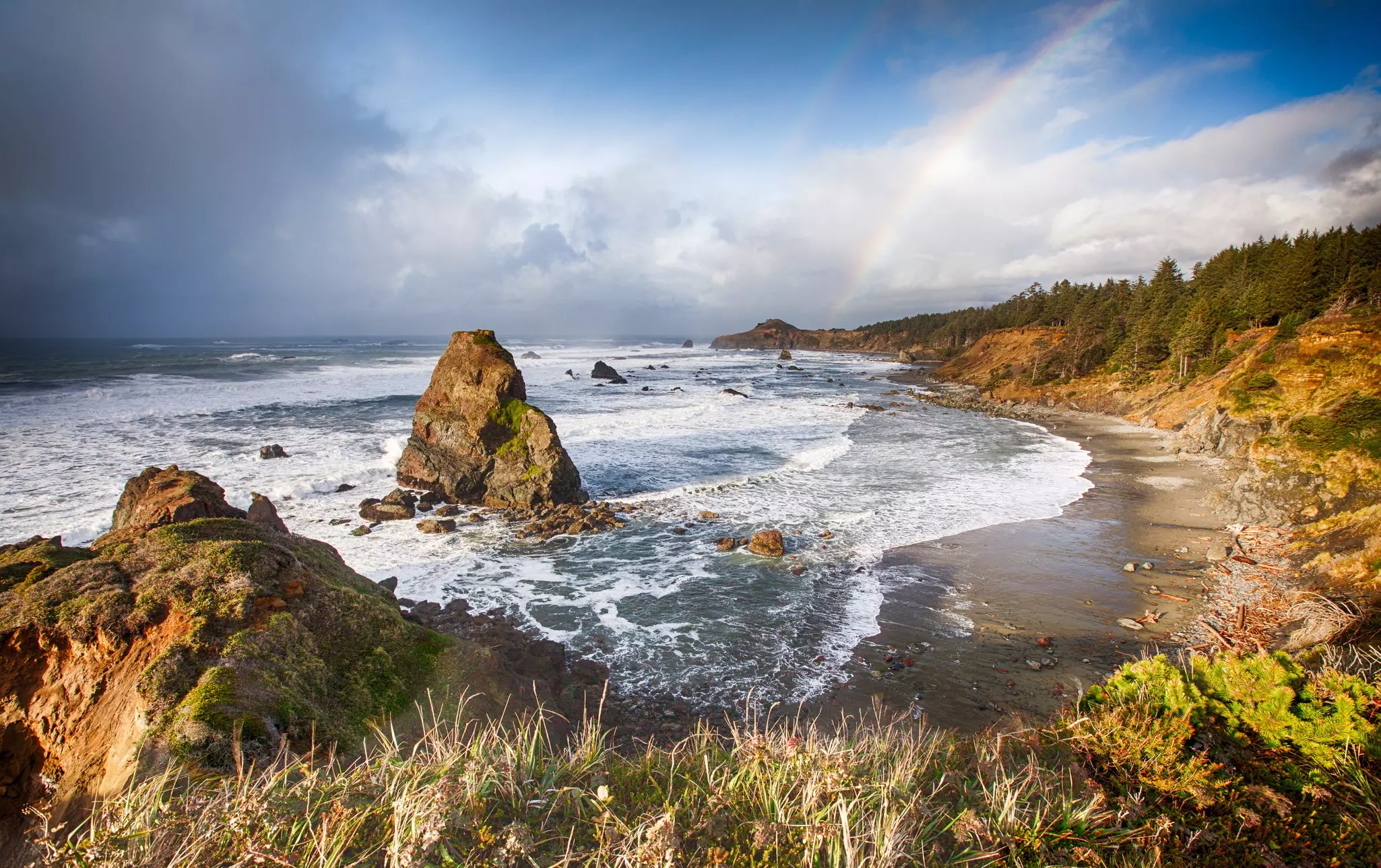 A beach backed by woodland. A double rainbow shines in the distance.
