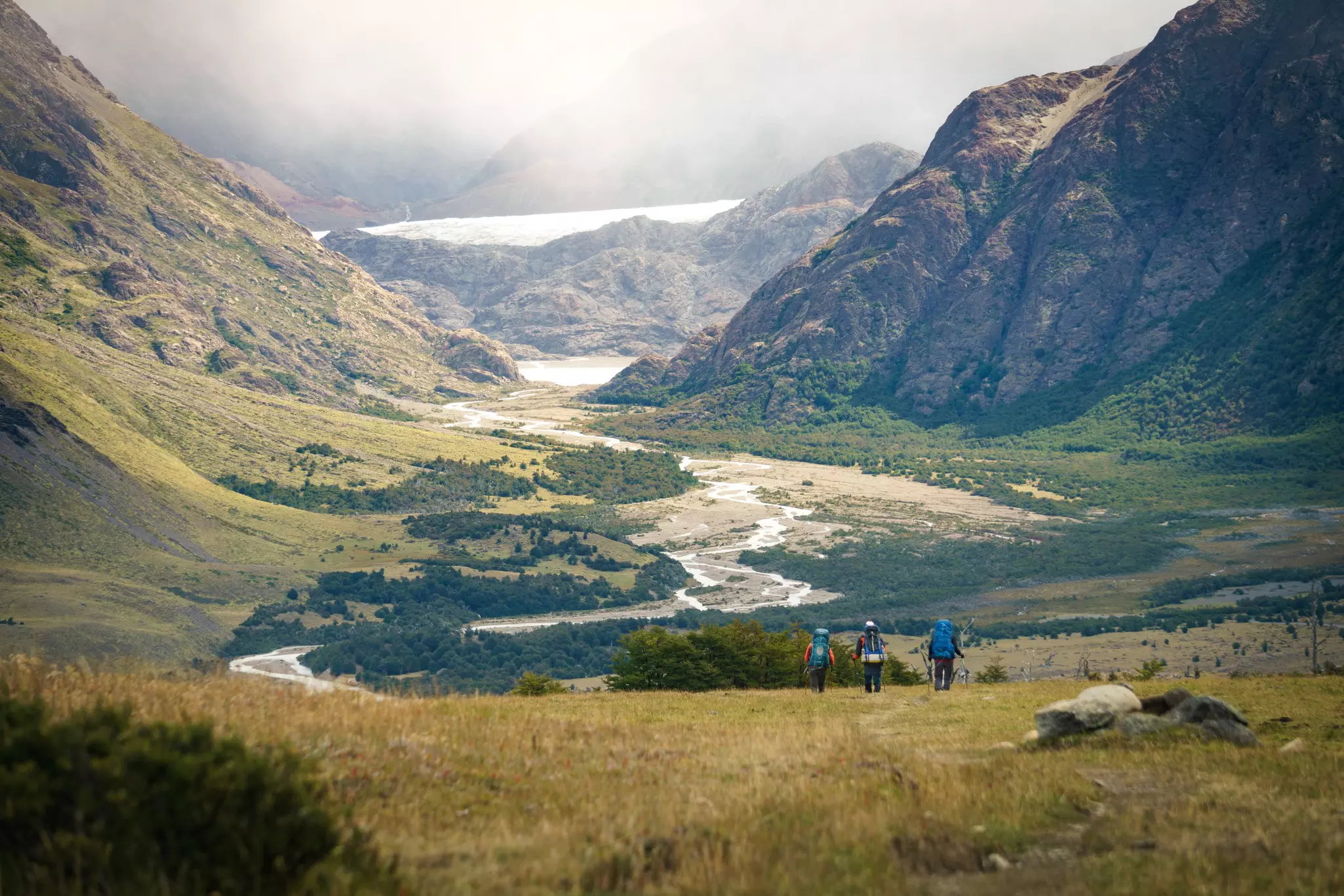 Three hikers with backpacks and poles walking through a valley with mountains in the distance on an overcast day.