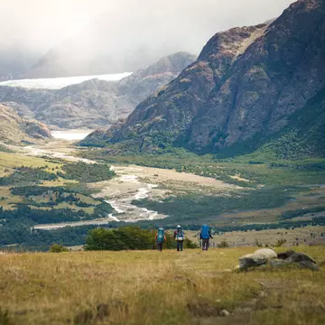 Hikers in a grassy valley approaching a snow-covered rocky peak.