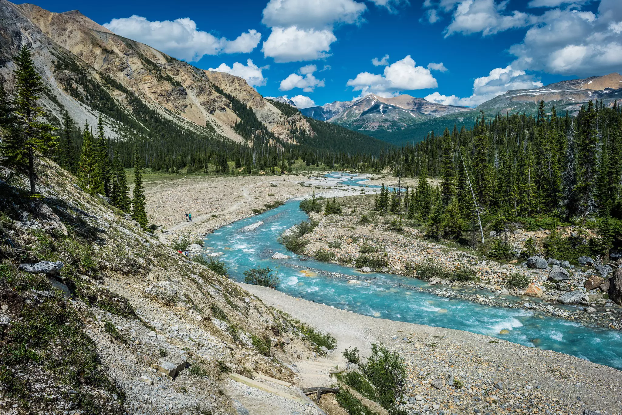 A rushing river runs through a valley; some people follow a hiking trail nearby