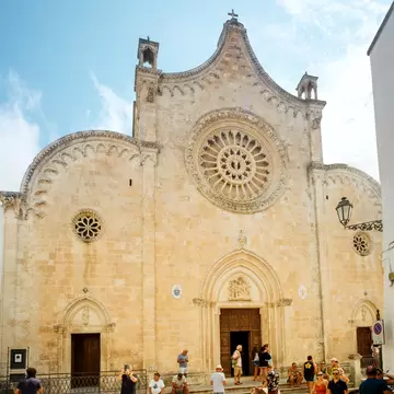 Ostuni Cathedral's pale stone facade with a large rose window above a carved Gothic doorway, visitors sitting on the steps