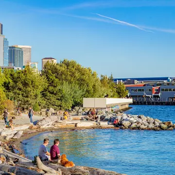 People sit on the sand of a small beach with a city skyline in the distance.