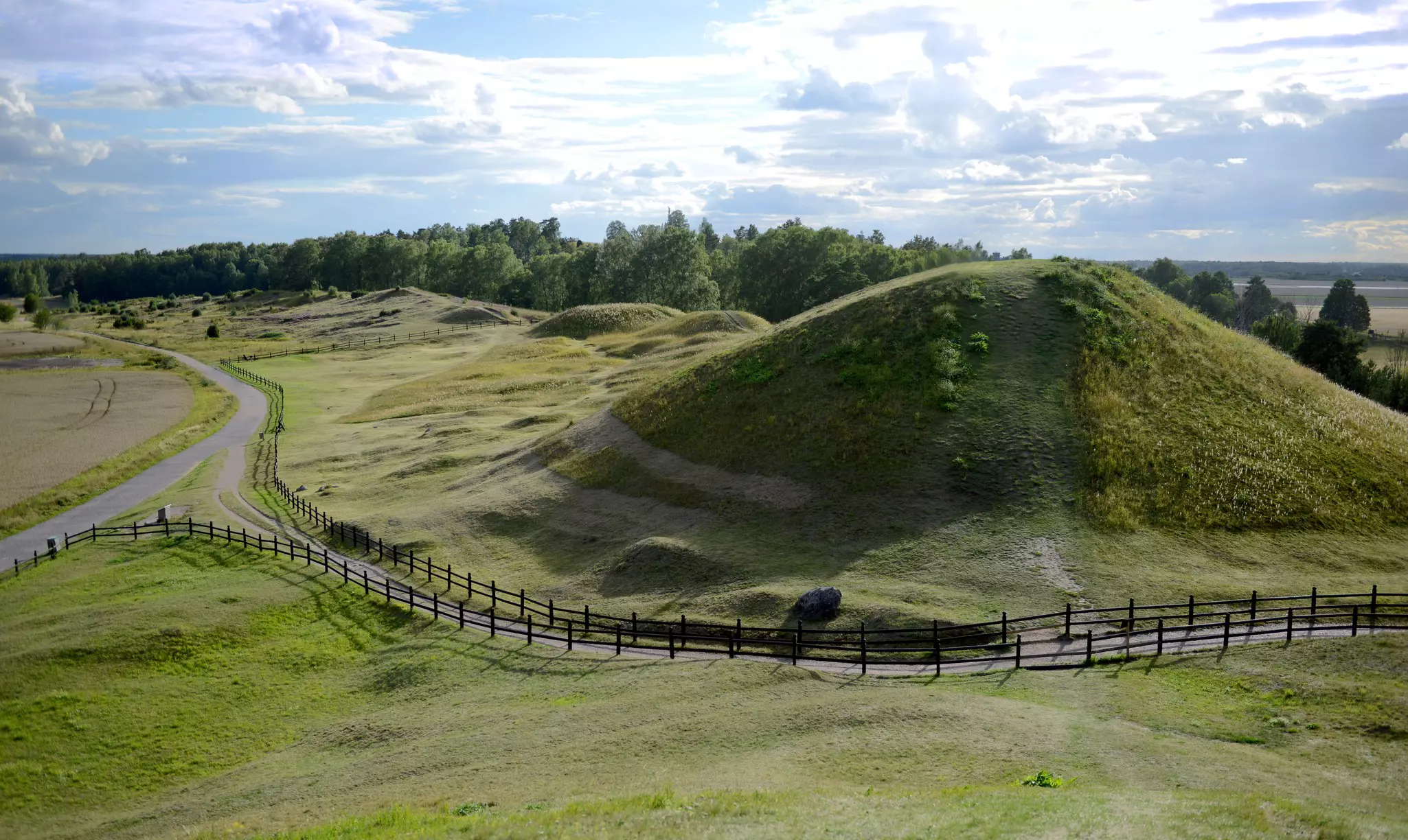 A large mound covered with grass is seen from afar, a fenced path snaking around it.