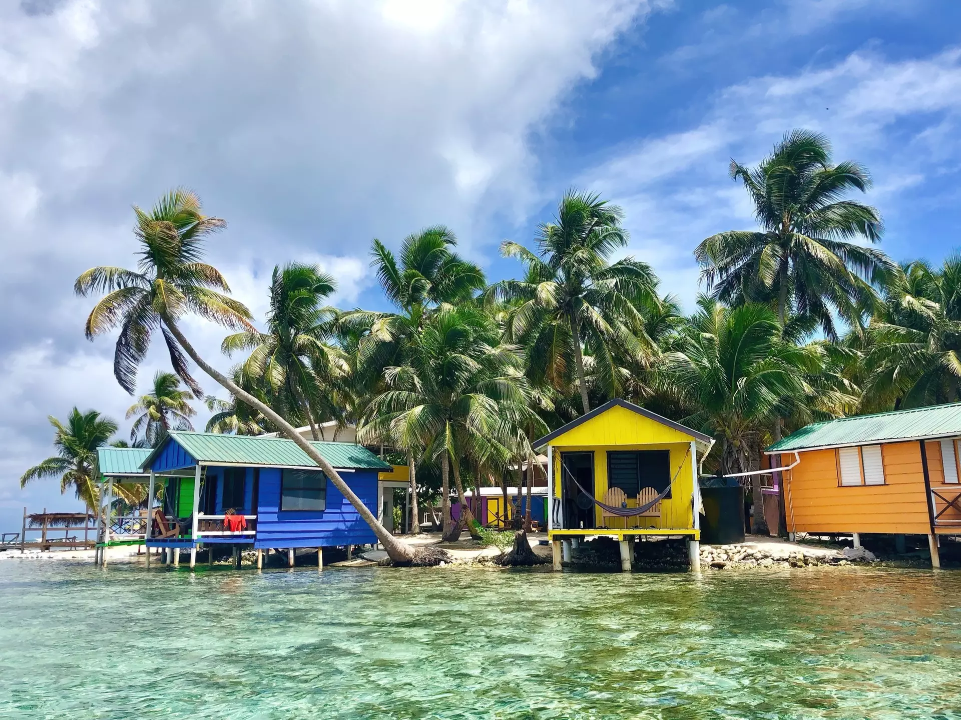 Colorful, small bungalows are built out over the clear water with palm trees behind them, Tobacco Caye, Belize
