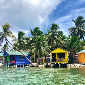 Colorful, small bungalows are built out over the clear water with palm trees behind them, Tobacco Caye, Belize