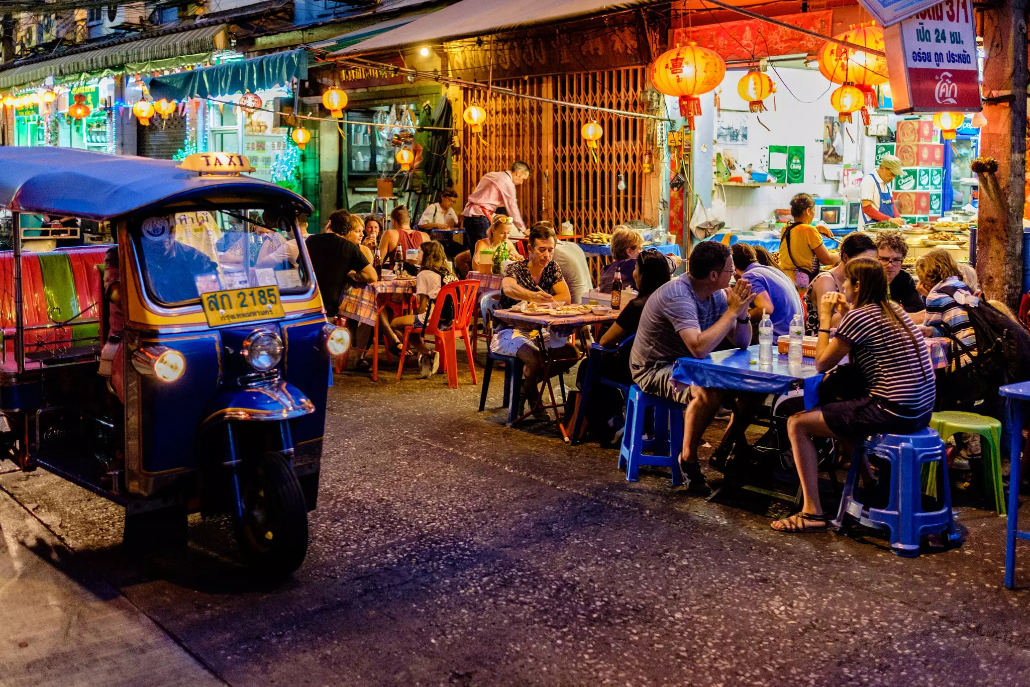 A busy night market with people sat on plastic chairs eating on the street outside a restaurant. A blue tuk-tuk is parked nearby.