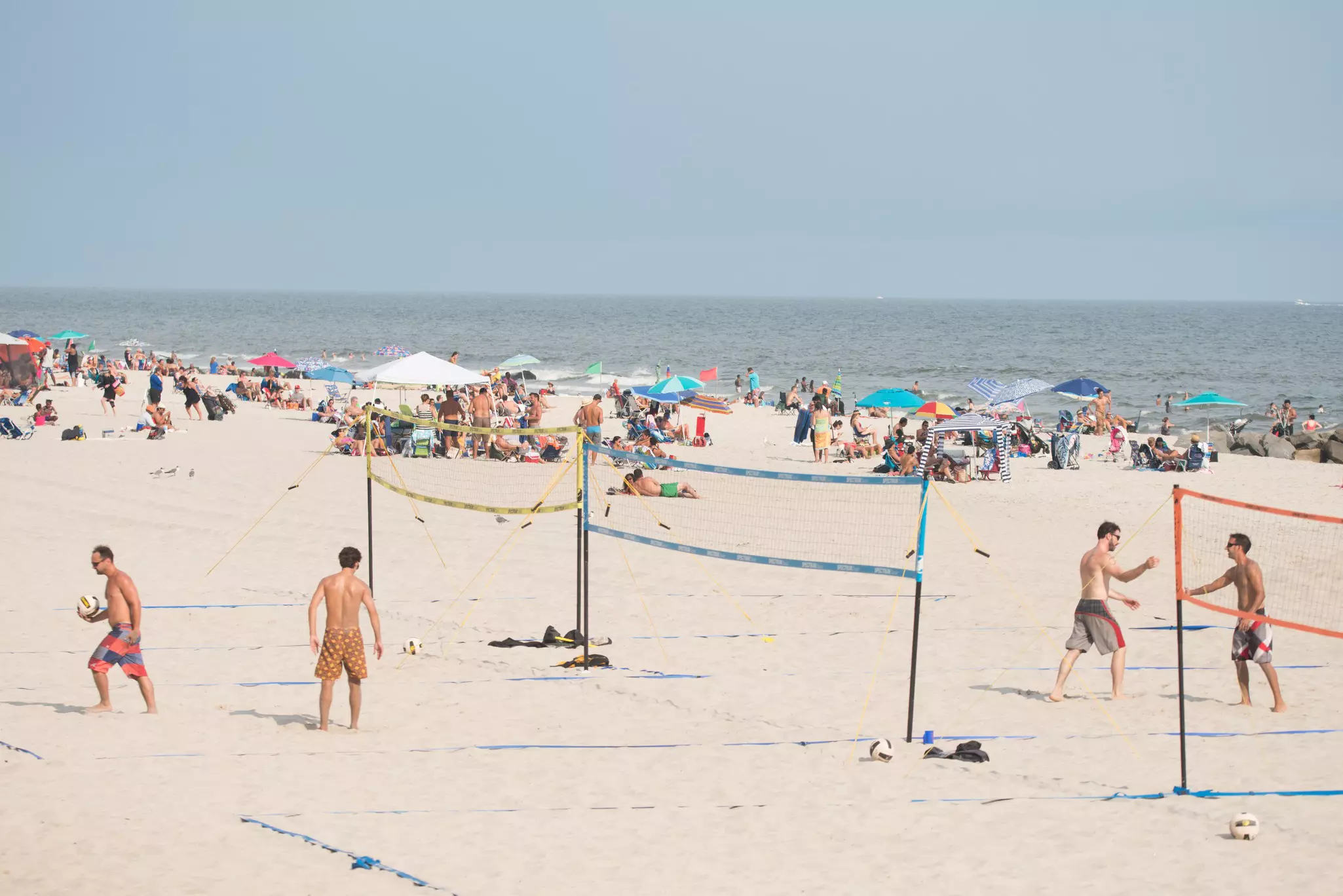 A large number of beach goers enjoying a beautiful sunny day with four men playing beach volleyball.