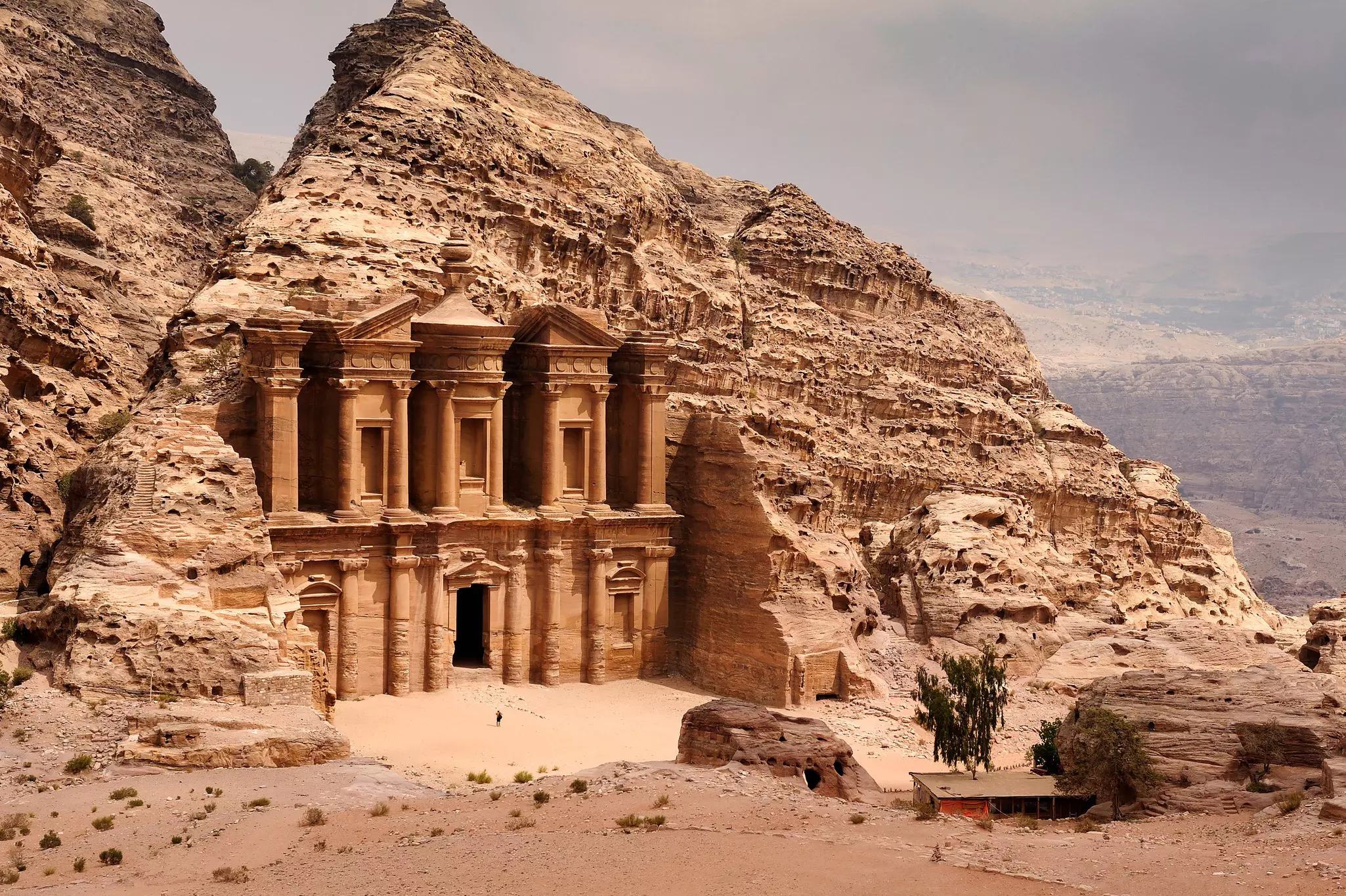 A person stands near a doorway to a massive building built and carved into stone in a hilly desert area of Jordan.