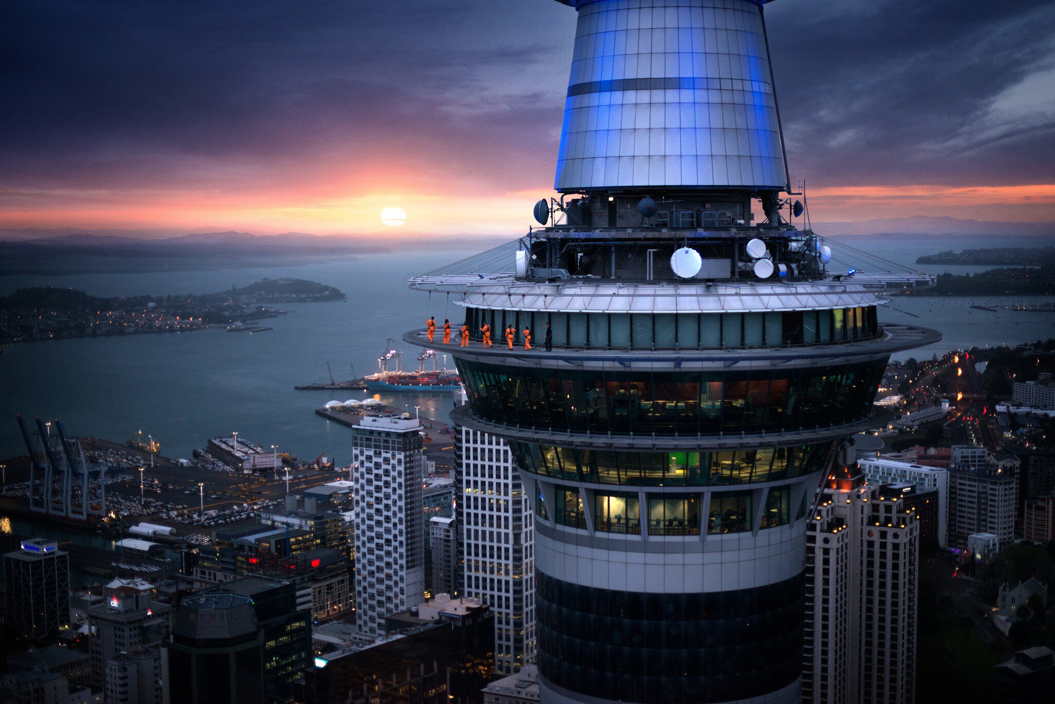 A group of thrill seekers in jumpsuits walk around the edge of the Sky Tower as the sun sets on the horizon in Auckland, New Zealand.