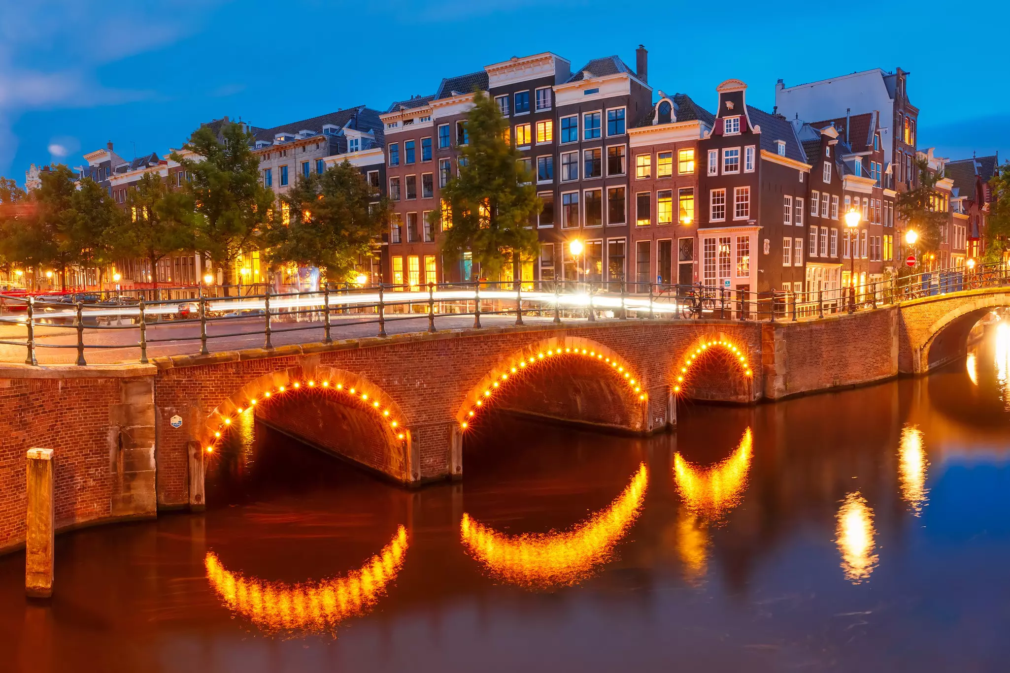 Nighttime view of canal with curved sets of lights reflected from a brick bridge above and lighted brick buildings in the distance.