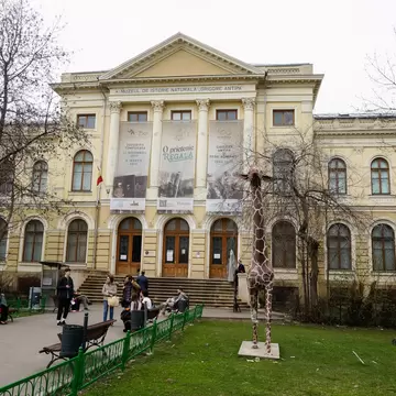 Yellow columned building hung with exhibition banners, bare trees, benches with visitors and a tall giraffe statue