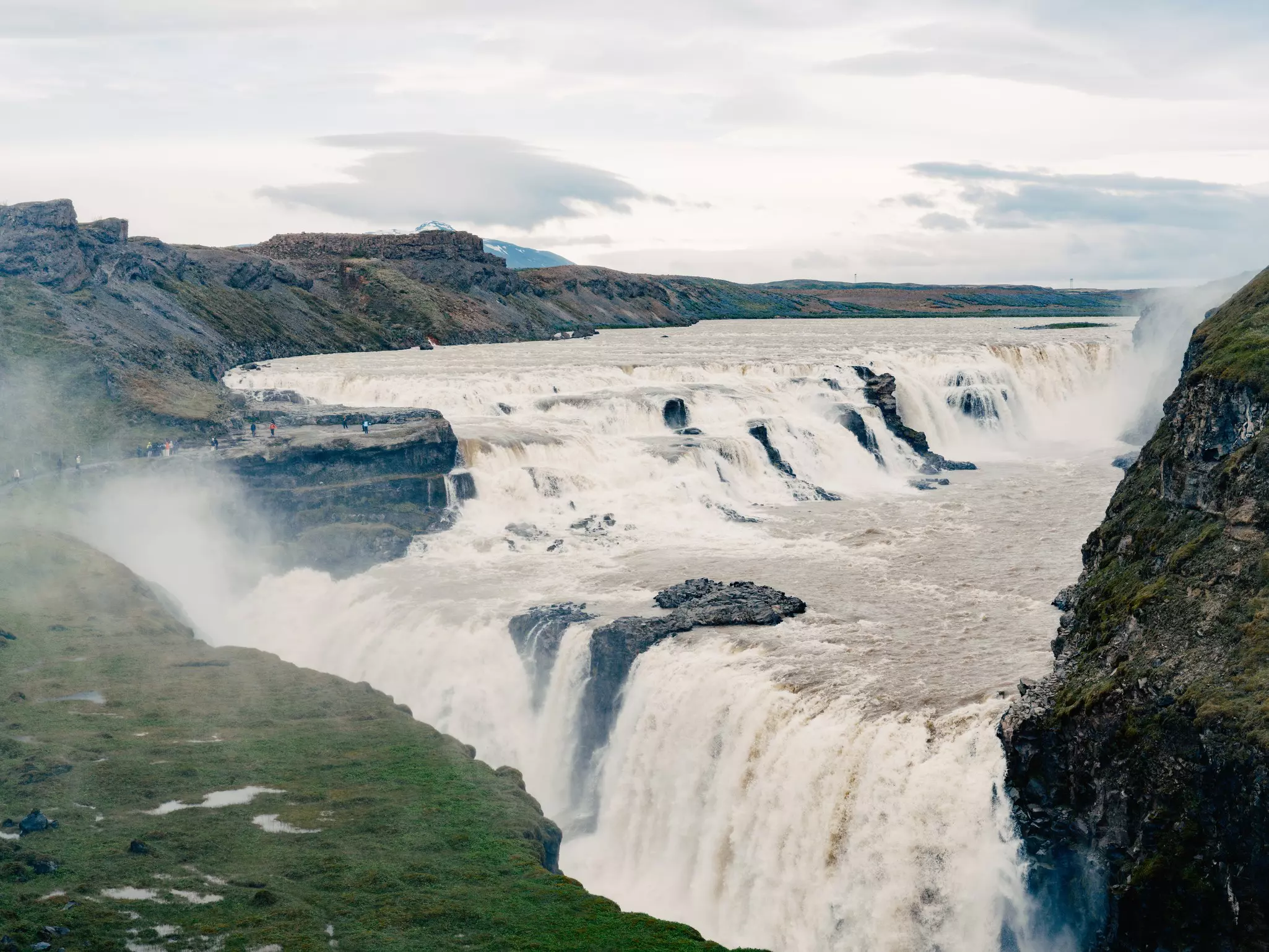 Aerial shot of wide body of water with numerous waterfalls spilling white water over rocky cliffs surrounded by low mountains. People walk on a path to the left in the far distance on an overcast day.