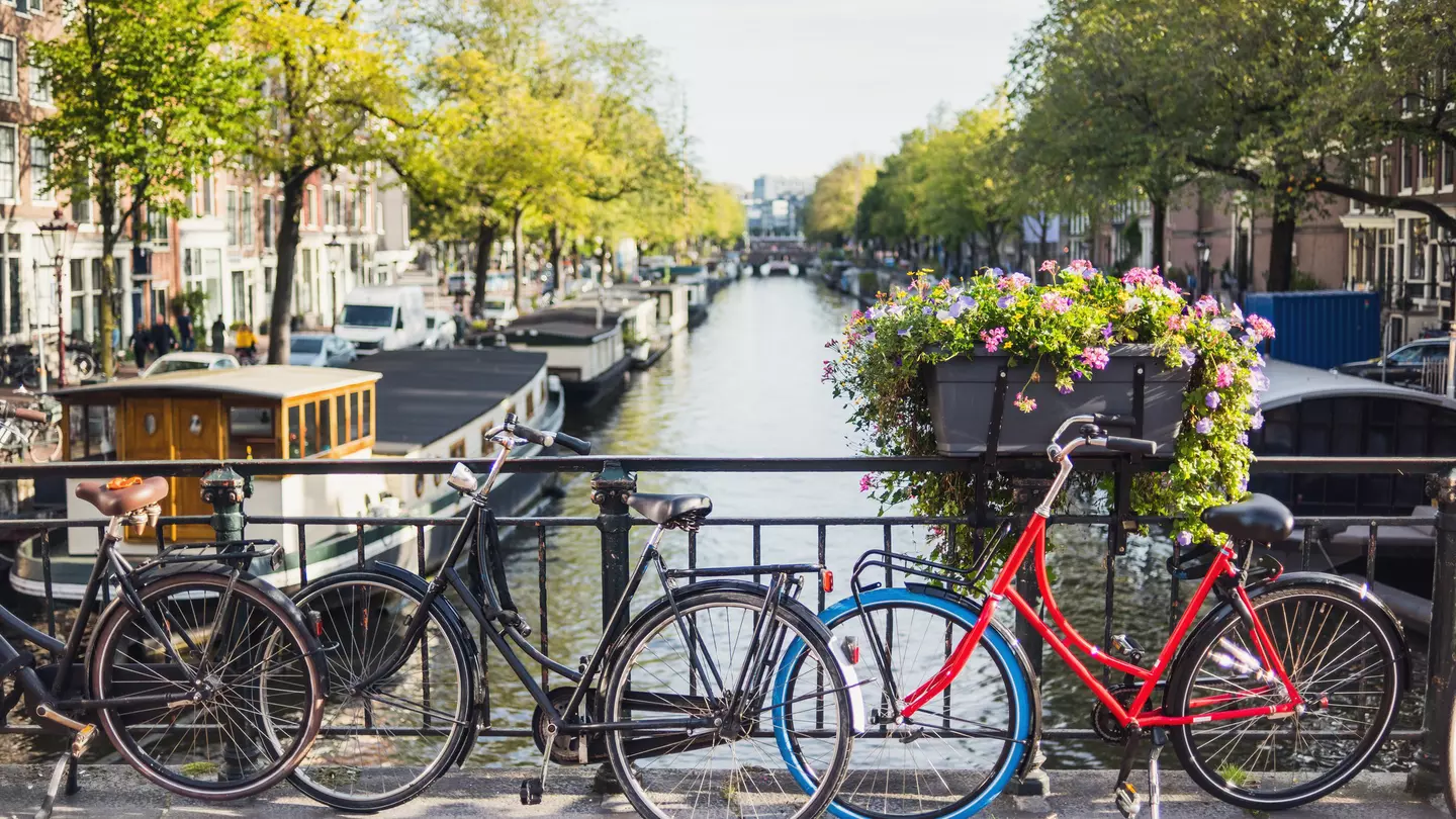 Three bikes locked to an iron fence above a canal with canal boats and brick buildings buildings in the distance.