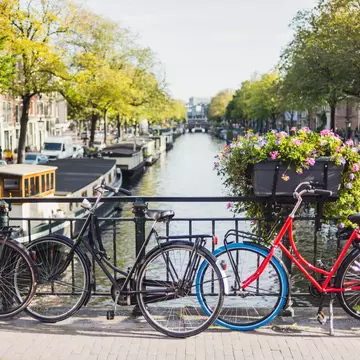 Three bikes locked to an iron fence above a canal with canal boats and brick buildings buildings in the distance.