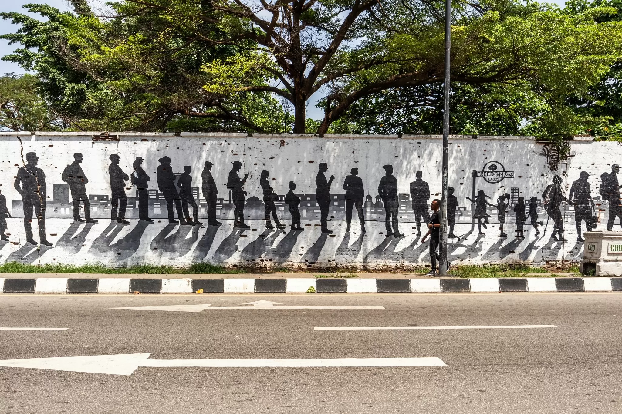 A long white wall painted with black silhouettes of people beside a road, shaded by trees at Freedom Park in Lagos.