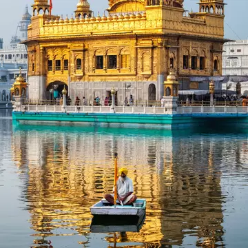A man in a yellow turban sits in a small boat on still water, with the gilded Golden Temple and its reflection behind him.