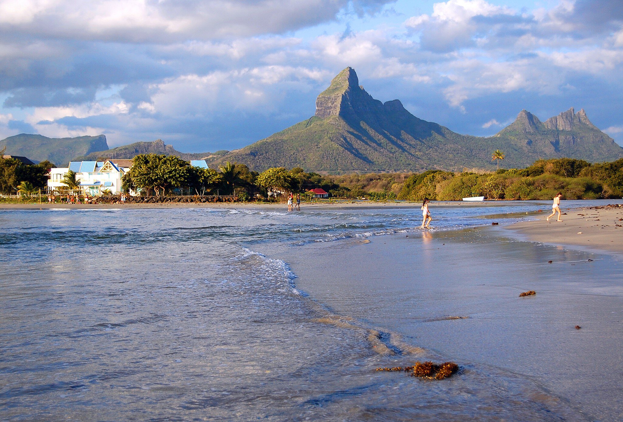Shallow tidal beach in low light with people at the waterline and a sharp volcanic peak behind the trees.