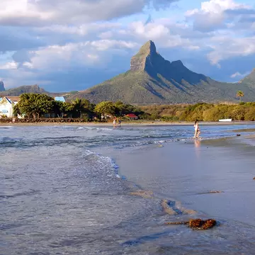 A shallow tidal beach in low light with people walking the waterline, a sharp volcanic peak and jagged ridge rising behind the treeline.
