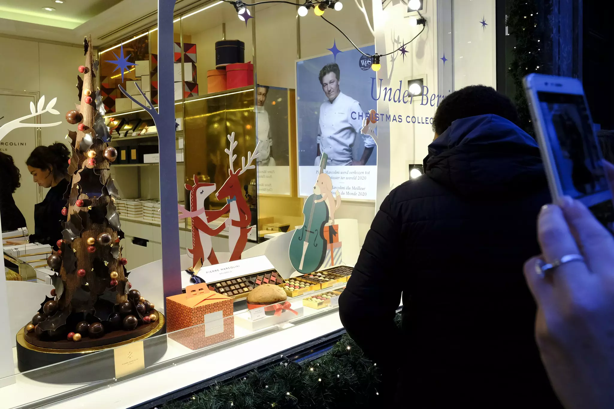 People stand outside a storefront looking at a window display that includes a chocolate tree decoration and boxes of chocolates.