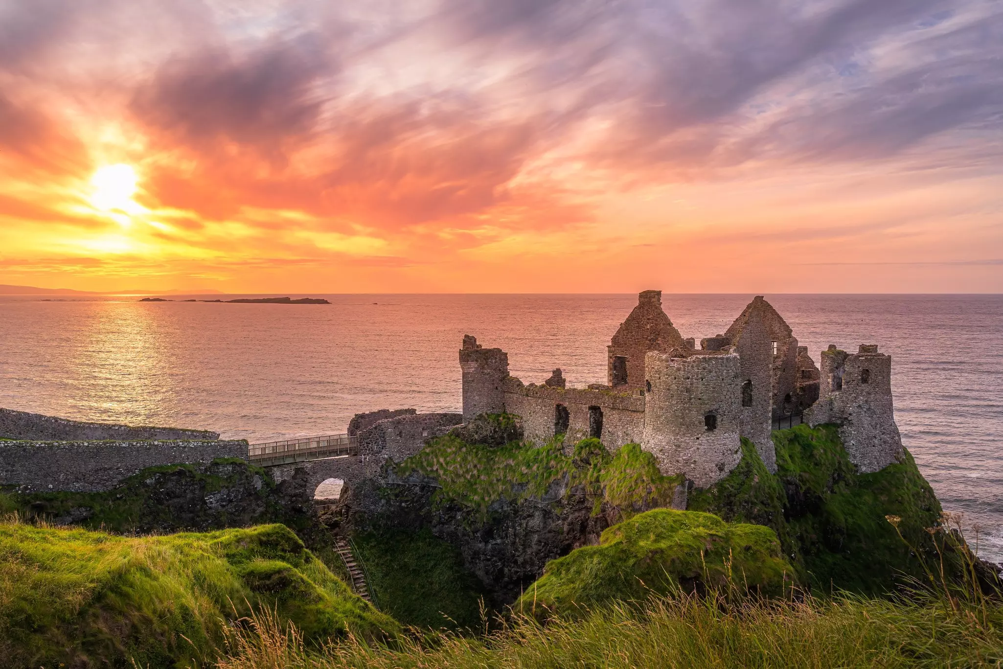 Ruins of a castle at the edge of a cliff. The sun sets in a colorful sky above it.