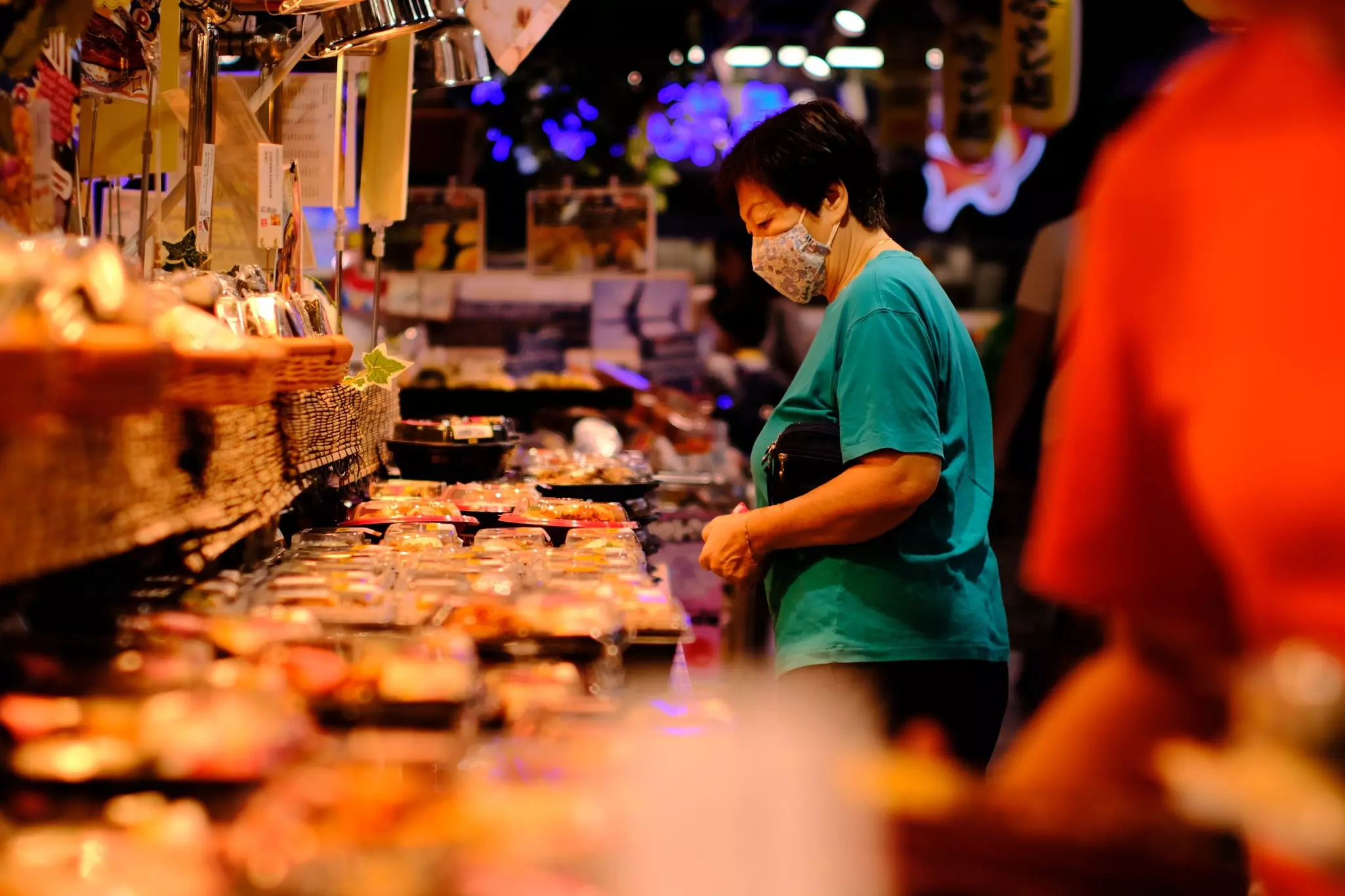 A woman in a mask looks at a variety of ready-to-eat foods for take out at a supermarket food hall.