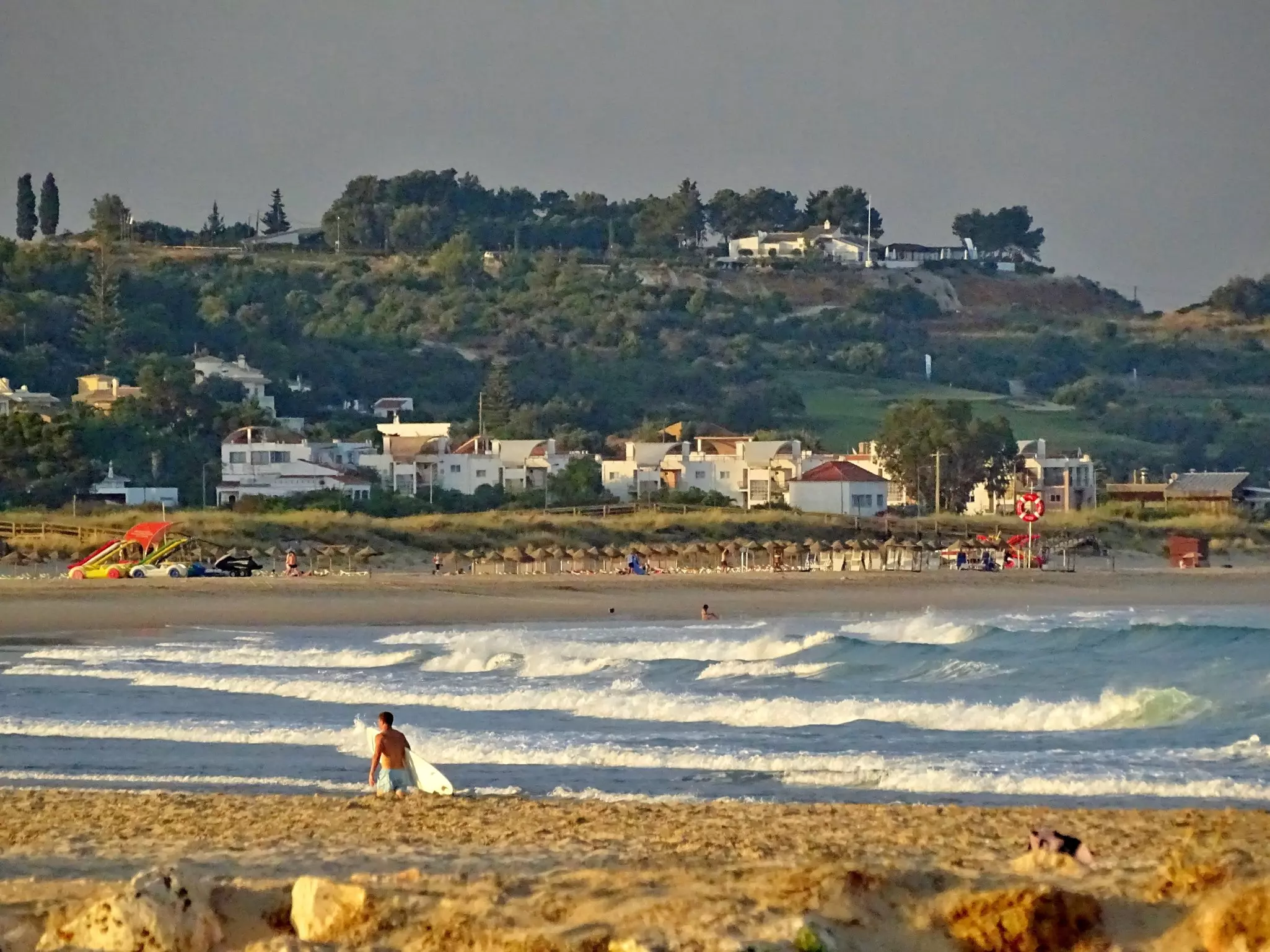 A surfer holding his board walks along a beach with whitecapped waves. Buildings are seen on the far shore.