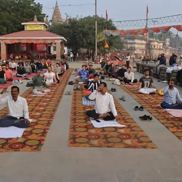 Rows of people sitting cross-legged on patterned rugs on a stone terrace, a small shrine and temple spire behind
